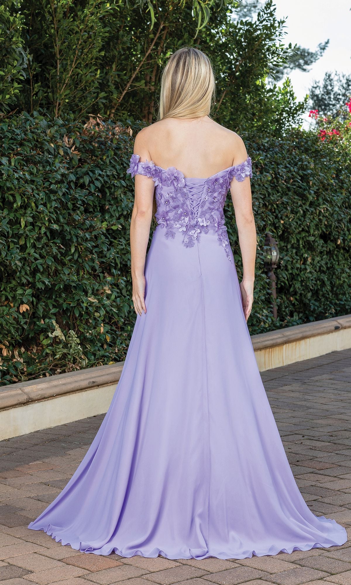A woman with blonde hair stands outdoors, shown from the back, wearing a lavender long corset-back prom dress with a beaded bodice.